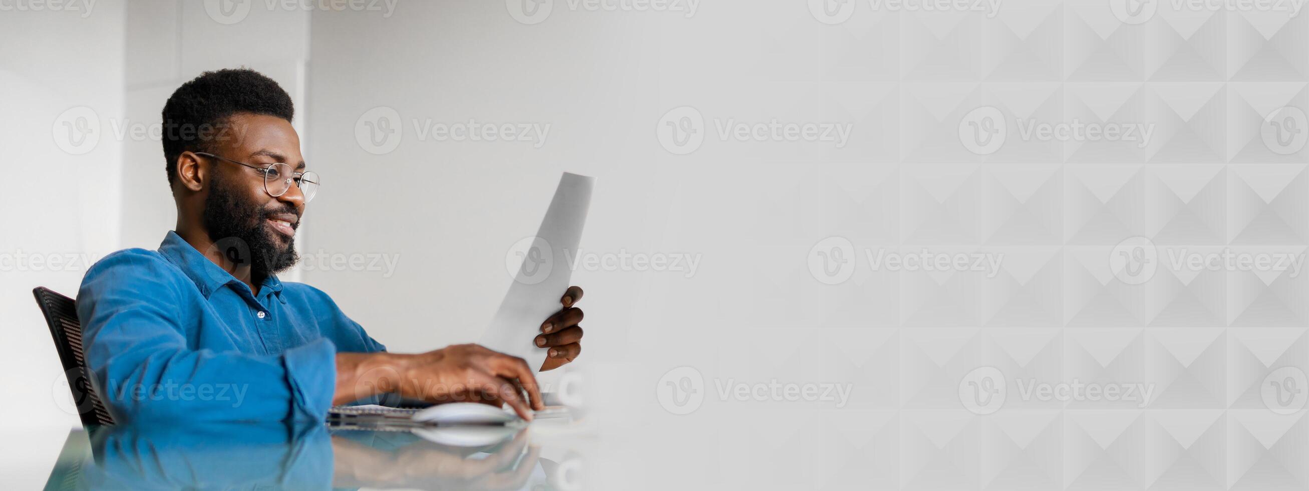 A young African American businessman reviews important documents while seated at a sleek desk. He is focused on his work in a bright, contemporary office environment, copy space photo