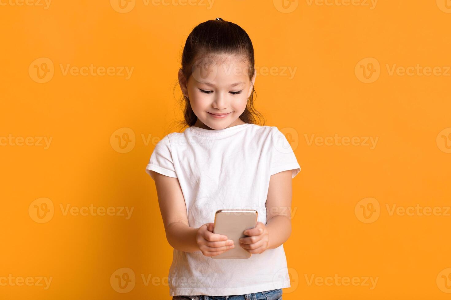 Mobile Games For Kids. Lovely asian girl using smartphone and smiling, sending message to parents or friends, browsing internet or watching cartoons, standing over pastel yellow wall, copy space photo