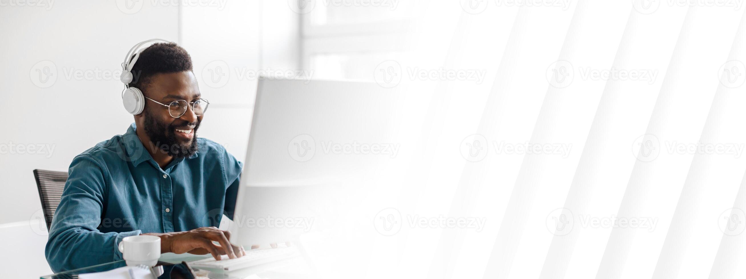 In a modern office environment, a young African American man, wearing headphones, is focused on his work at a computer. His expression reflects determination and success, copy space photo