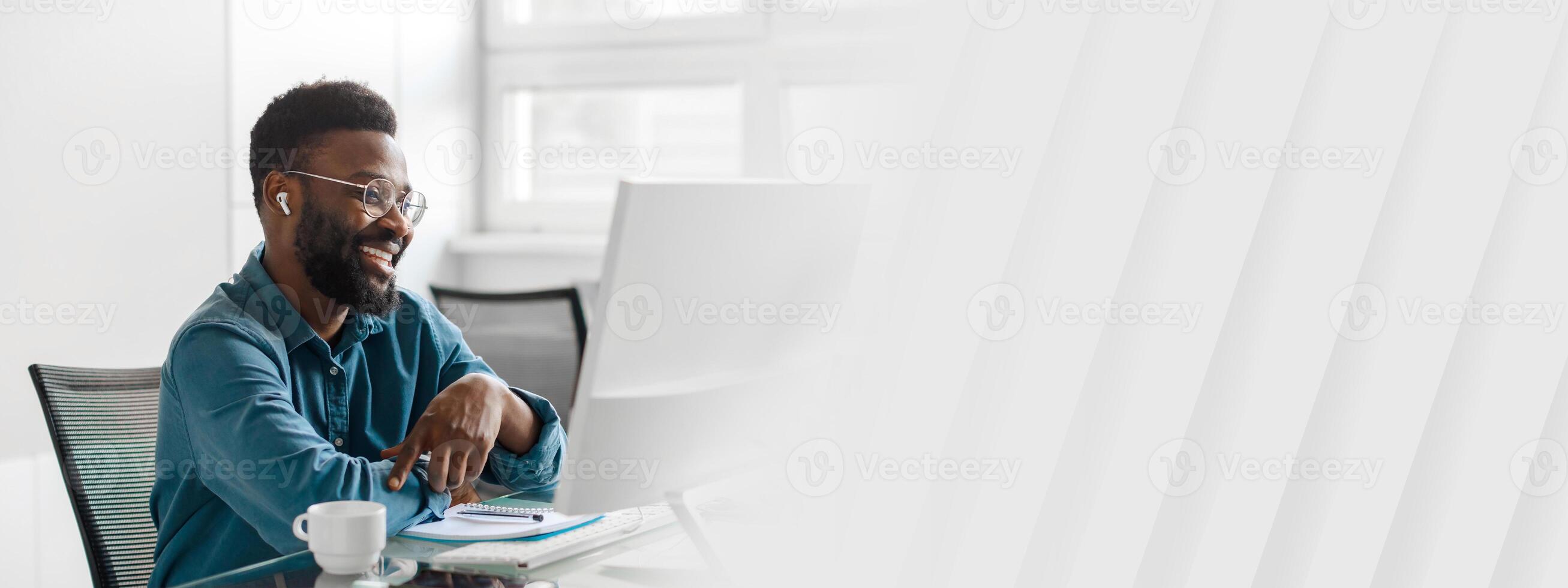 A young African American entrepreneur sits at a desk in a bright office. He smiles while engaged in work, surrounded by a clean and modern space, ready for success, copy space photo