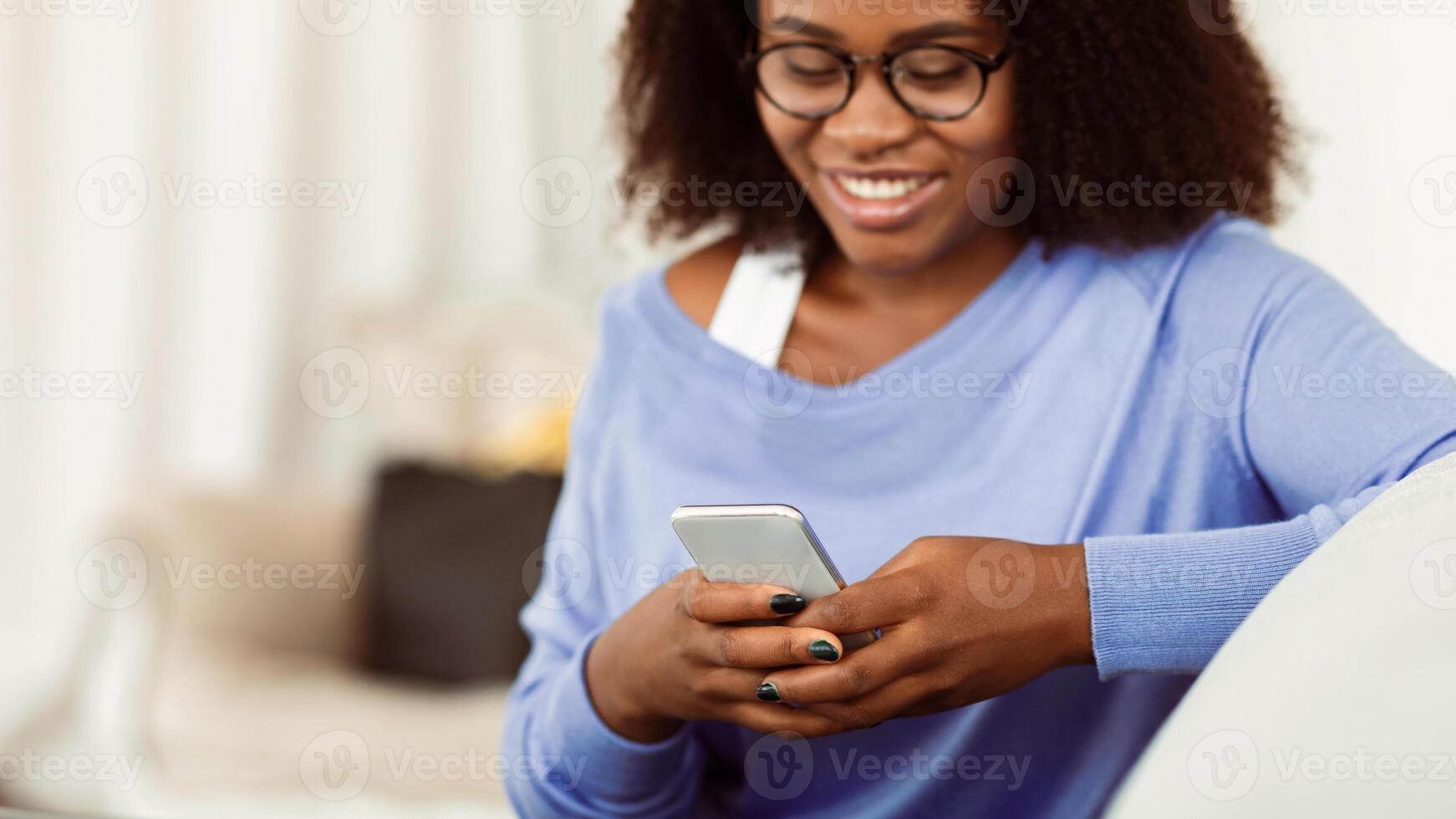 Scrolling News Feed. Closeup of african american smiling lady holding mobile phone, typing sms message, sitting on couch. Communication Concept. Black woman browsing internet, surfing web, using app photo