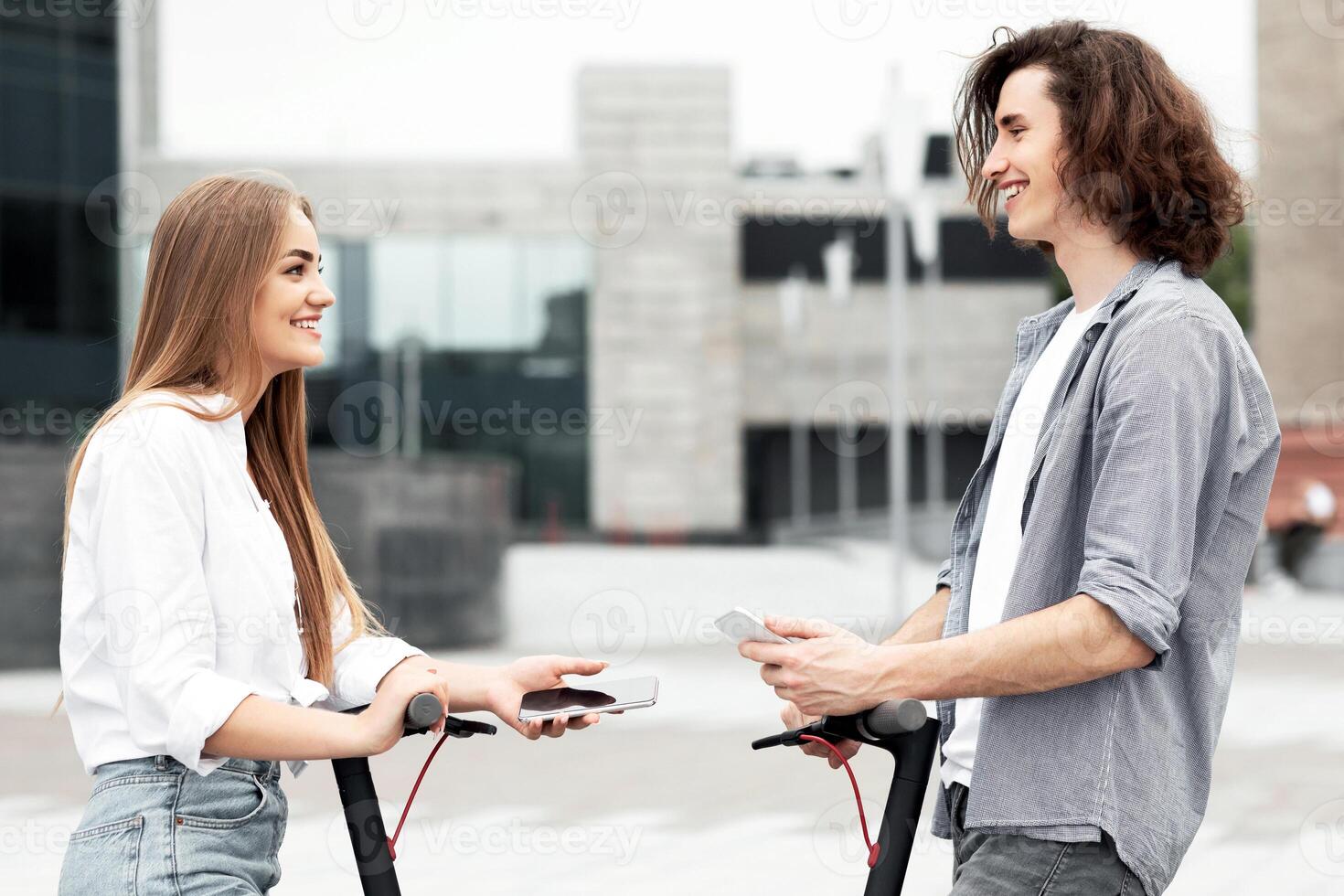 Two teenagers using e vehicle rent service with smart phones in urban city street, side view, blurred background photo