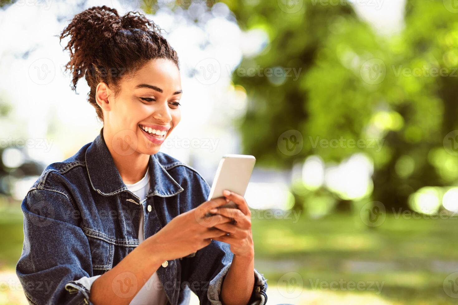 Happy Black Girl Using Mobile Phone App Browsing Internet Sitting In Park Outside. Empty Space For Text photo