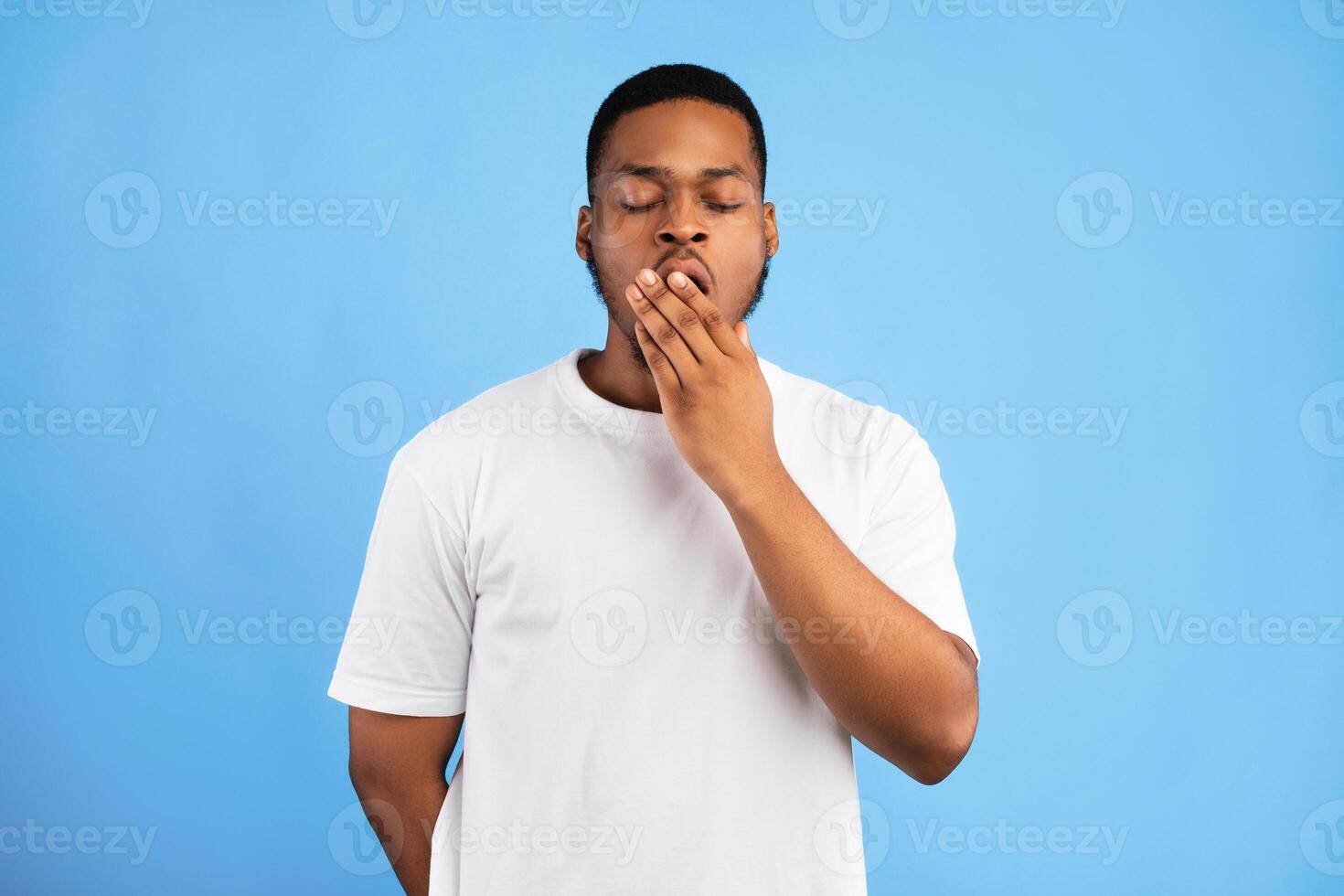Boredom. Sleepy Bored African American Guy Yawning With Eyes Closed Standing Over Blue Background. Studio Shot photo