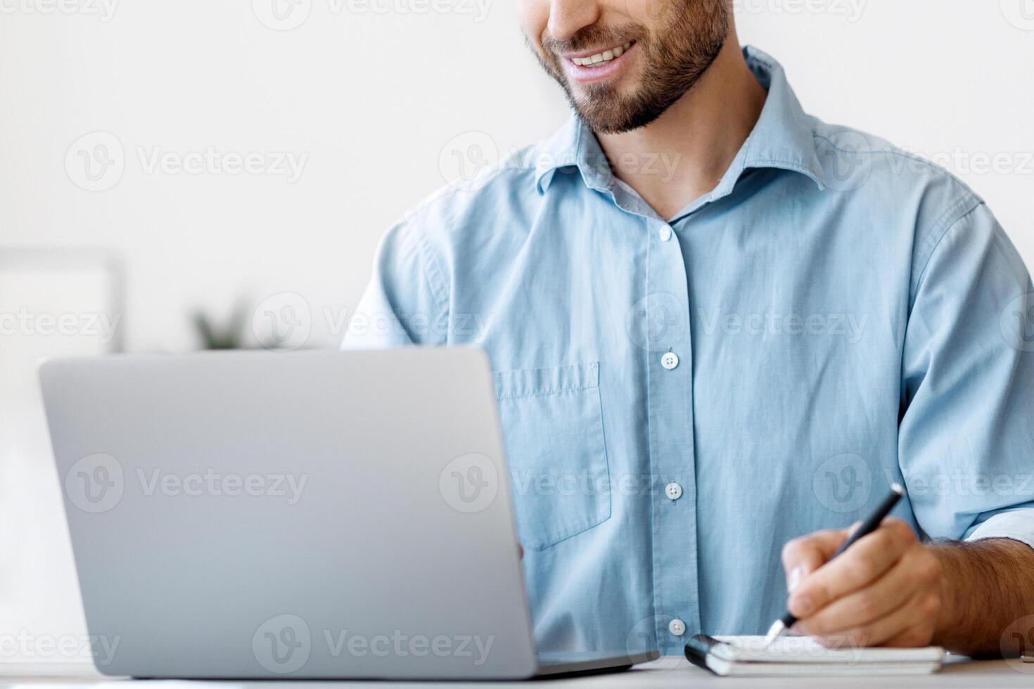 Millennial male entrepreneur using laptop and taking notes at workplace in modern office, looking at computer screen and writing something to notepad, sitting at desk, planning budget, free space photo