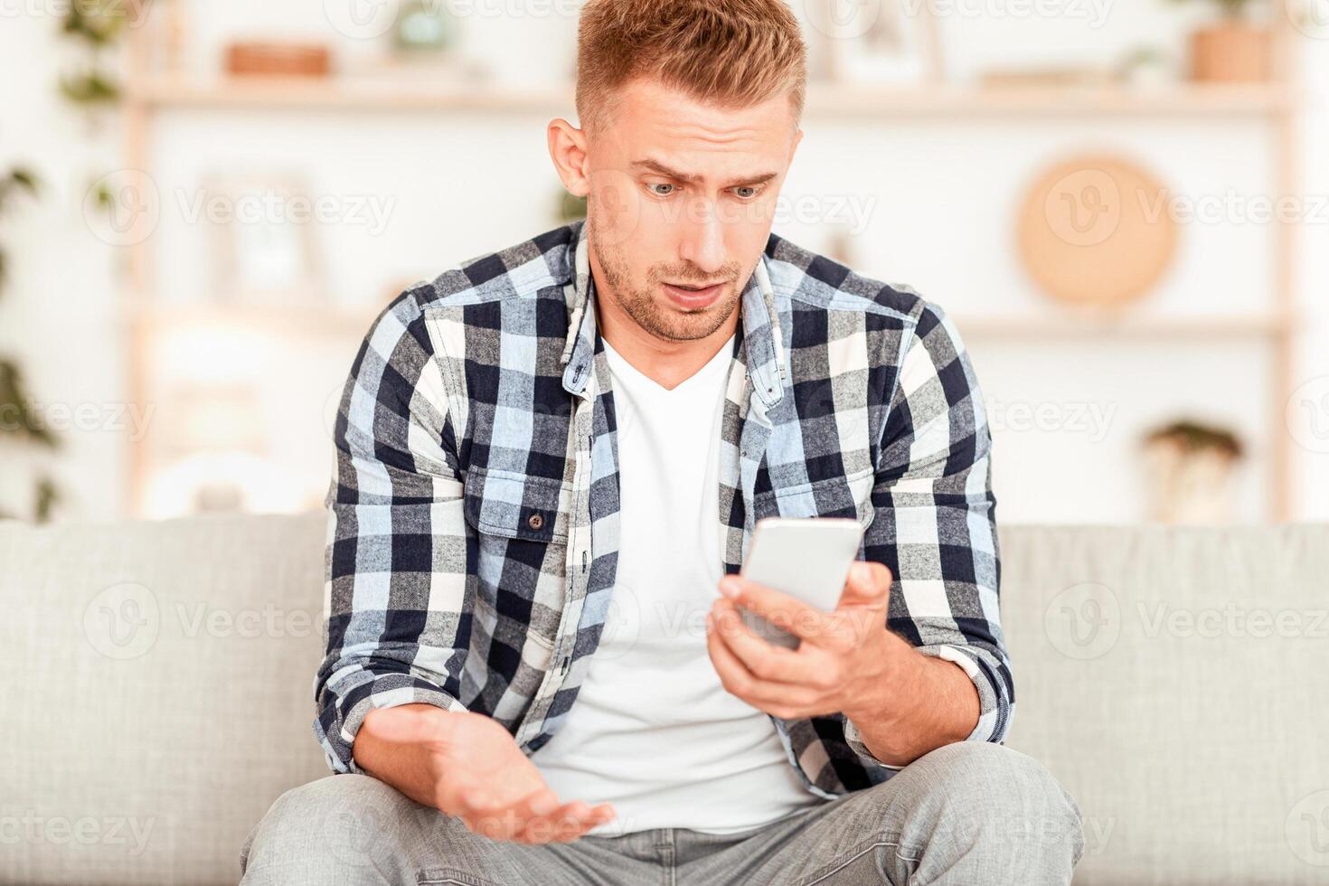 Bad News Concept. Frontal portrait of surprised shocked man looking at smartphone, disappointed by content from social media or message, sitting on the couch at home. Confused guy using mobile phone photo