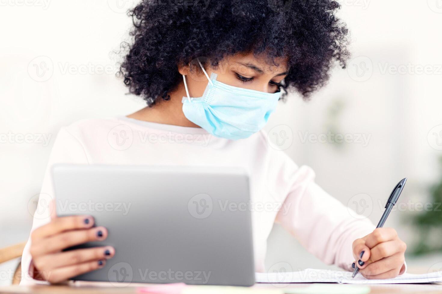 Black Schoolgirl Learning Using Digital Tablet And Wearing Protective Face Mask Sitting At Table Indoor. Virus Protection At School Concept photo