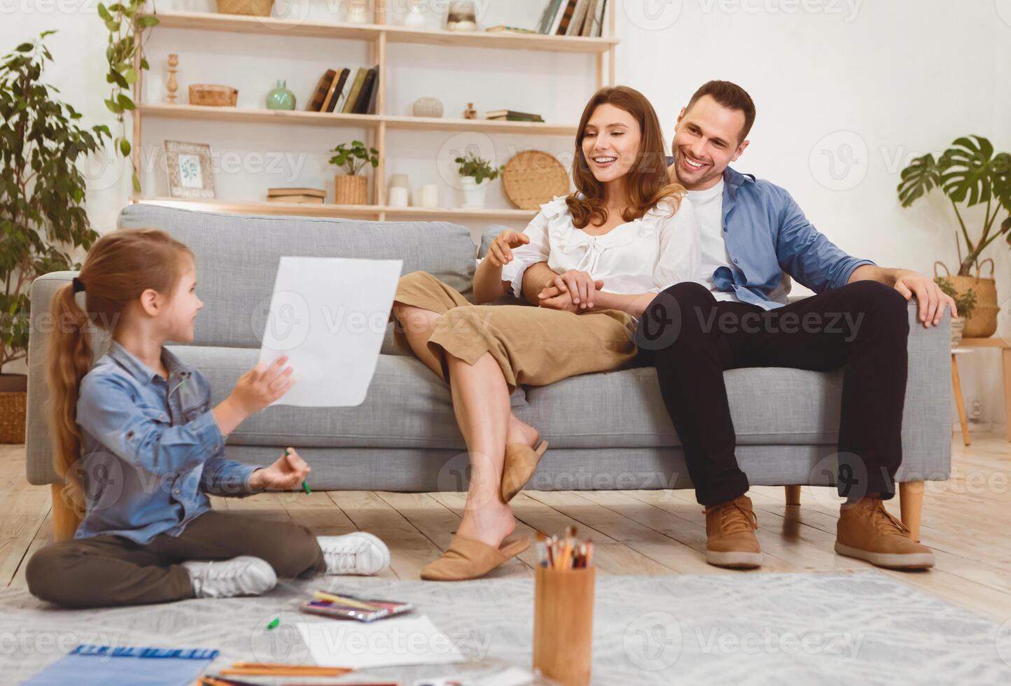 Weekend. Little Daughter Drawing Sitting On Floor, Showing Her Sketch To Parents Spending Time Together In Living Room At Home. photo