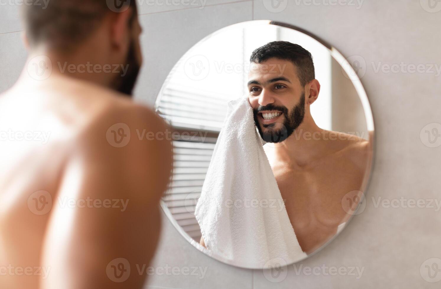 Male Facial Skincare Routine Concept. Over The Shoulder View Of Happy Shirtless Young Handsome Man Wiping His Face With White Towel Standing In Modern Bathroom Looking At Reflection In The Mirror photo