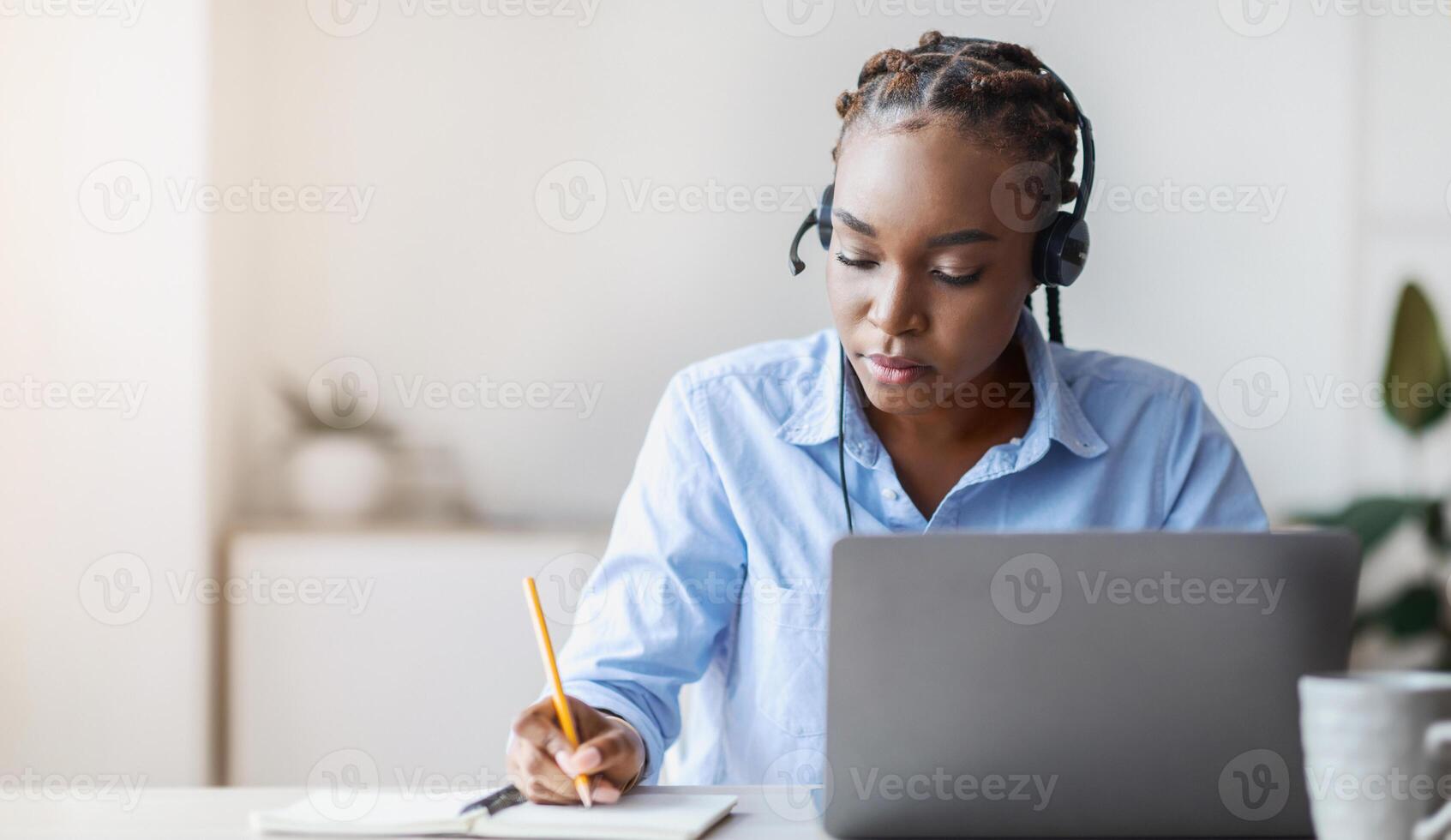 Online Training. Young black female employee in headset watching webinar on laptop and taking notes, sitting at desk in modern office, enjoying modern education and distance learning, copy space photo