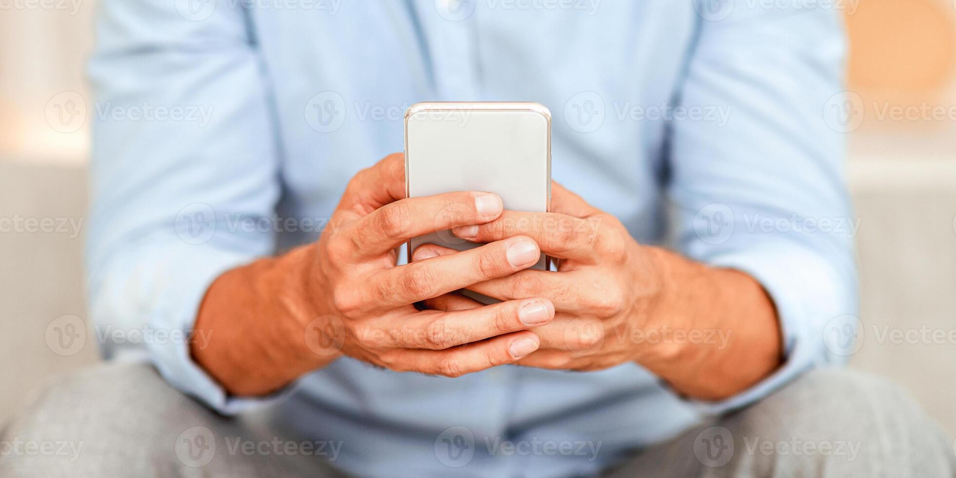 Gadget Addiction Concept. Closeup of unrecognizable guy holding mobile phone, typing sms message, sitting on couch, blurred background. Adult man browsing internet, surfing web, using app photo