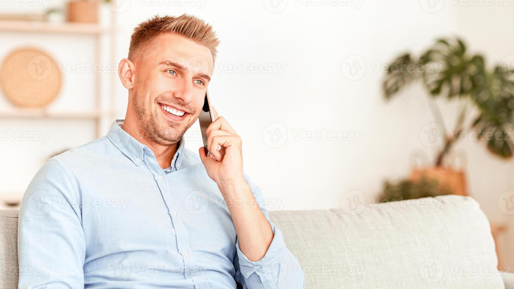 Mobile Communication Concept. Cheerful man making phone call, talking with his friend or family, sitting on the couch in living room. Confident adult calling, resting on sofa, free space photo