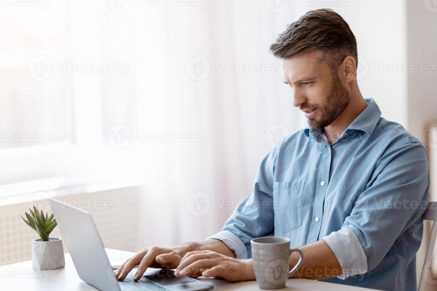 Unemployed man scrolling job search website at home using laptop computer, handsome male freelancer working remotely at desk near window, typing on keyboard, answering emails, copy space photo