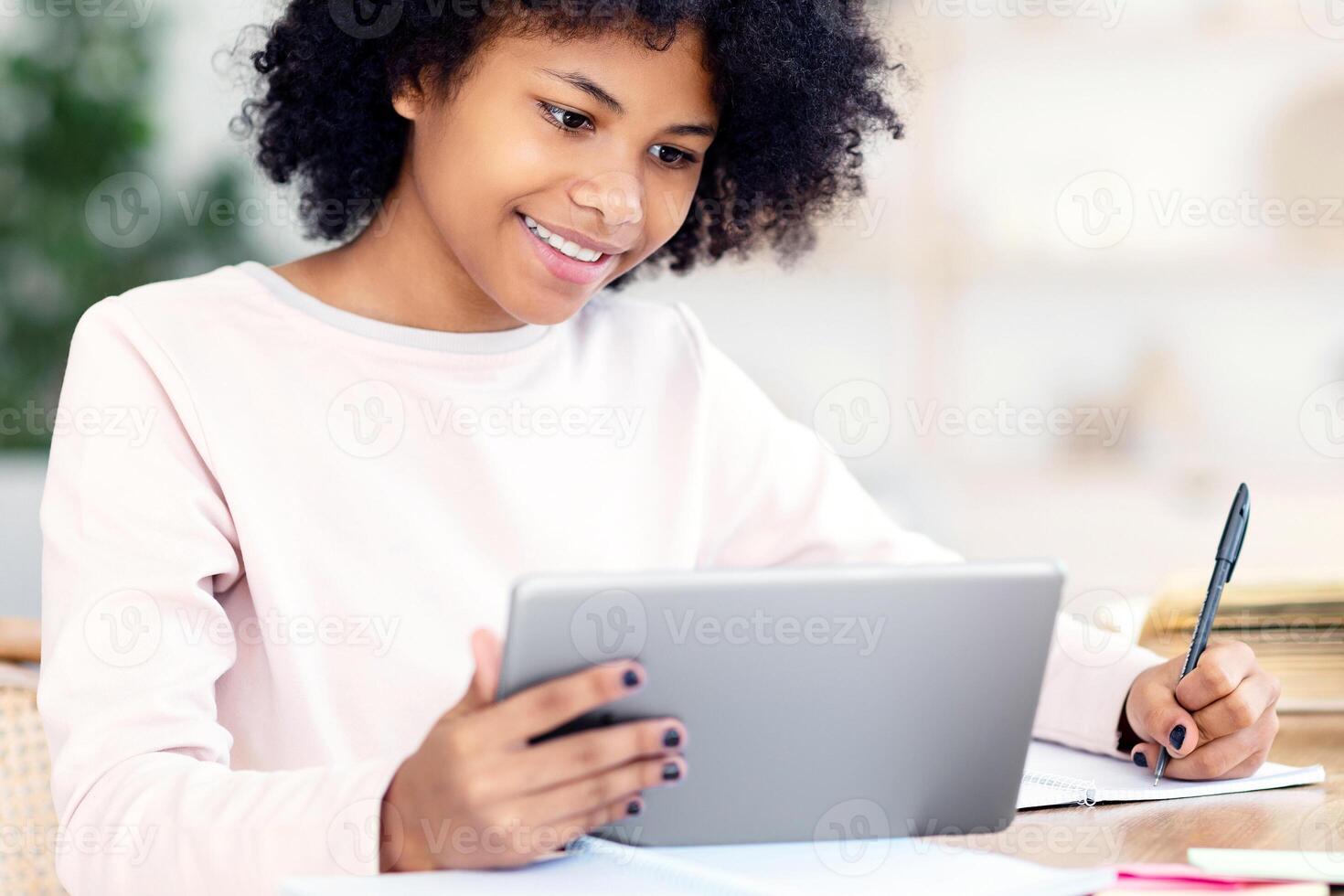 Cheerful Kid Girl With Digital Tablet Doing Homework Online Sitting At Desk At Home. Distant E-Learning Concept photo