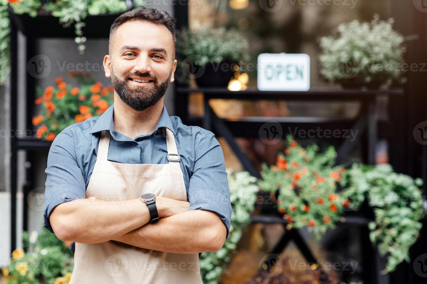 Modern business and new normal after covid-19 outbreak, open store and meeting client, outdoor. Smiling confident man in apron with crossed arms near flower shop with plants in pots and sign is open photo