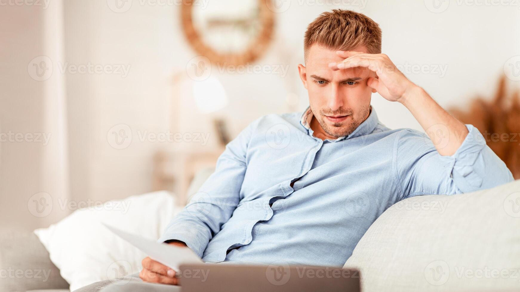 Foused man working from home, sitting on sofa, holding document and looking at personal computer photo