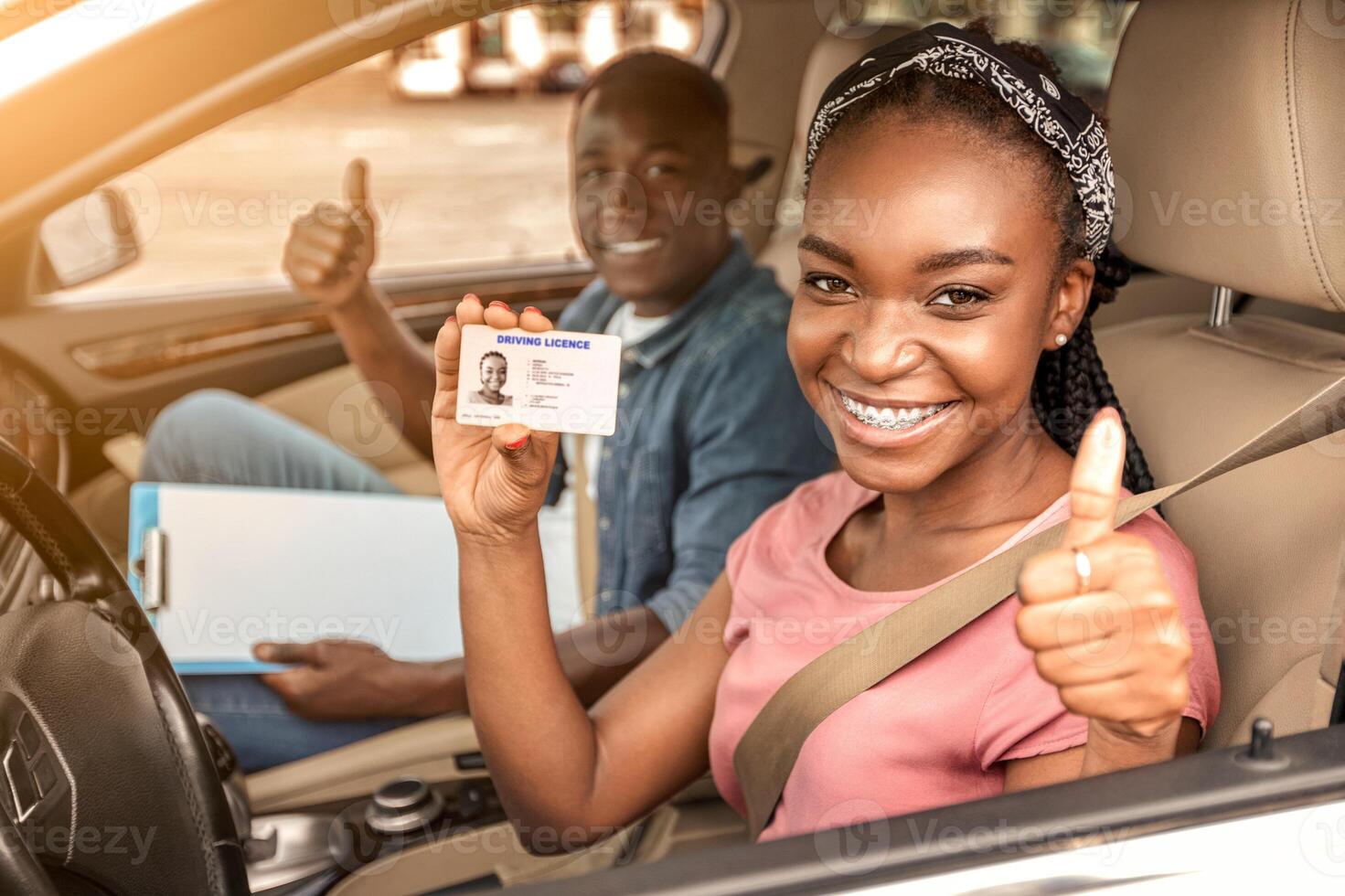 Happy african girl with driving license and her instructor showing thumb up inside car, woman passed driving test photo
