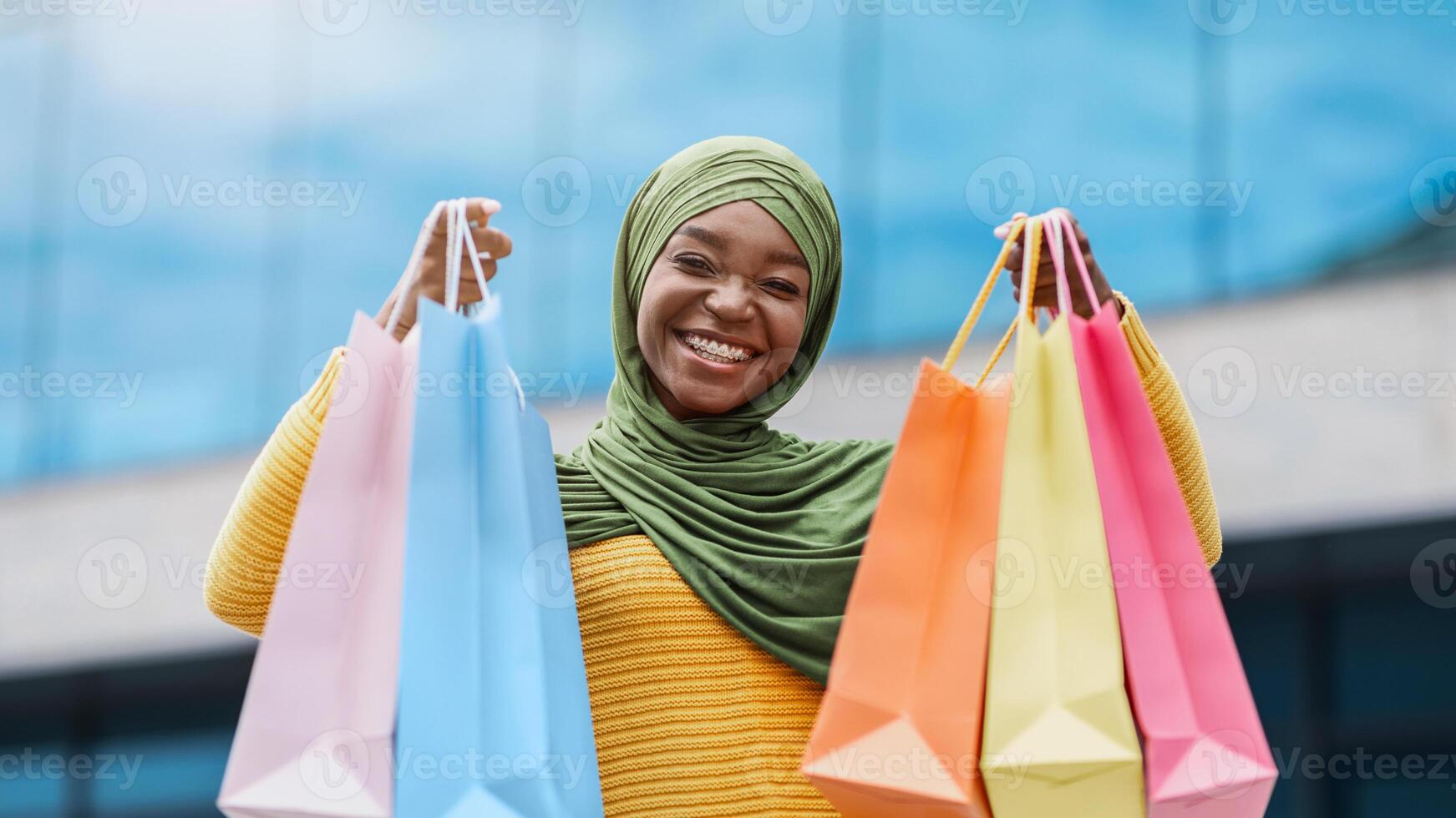 Happy black muslim woman in hijab with lots of shopping bags posing outdoors, demonstrating purchases to camera, enjoying seasonal sales and discounts, portrait of islamic shopaholic lady, copy space photo