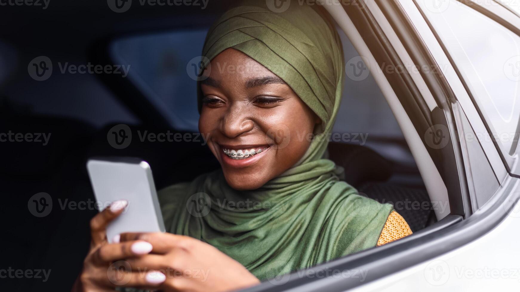Smiling Black Muslim Lady In Hijab Using Smartphone While Riding Car On Backseat, Browsing Internet Or Texting With Friends, African Woman Enjoying Online Communication, Closeup Shot Through Window photo