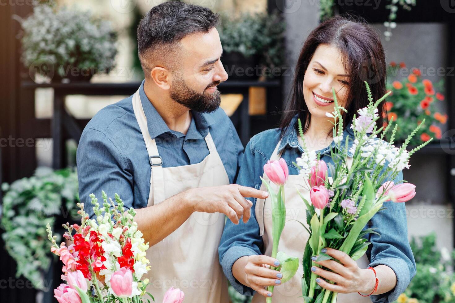 Florist teaches assistant to make bouquet for client online order. Millennial man helps to lady in apron to make composition at counter at flower shop with diverse plants in pots and vases outdoor photo