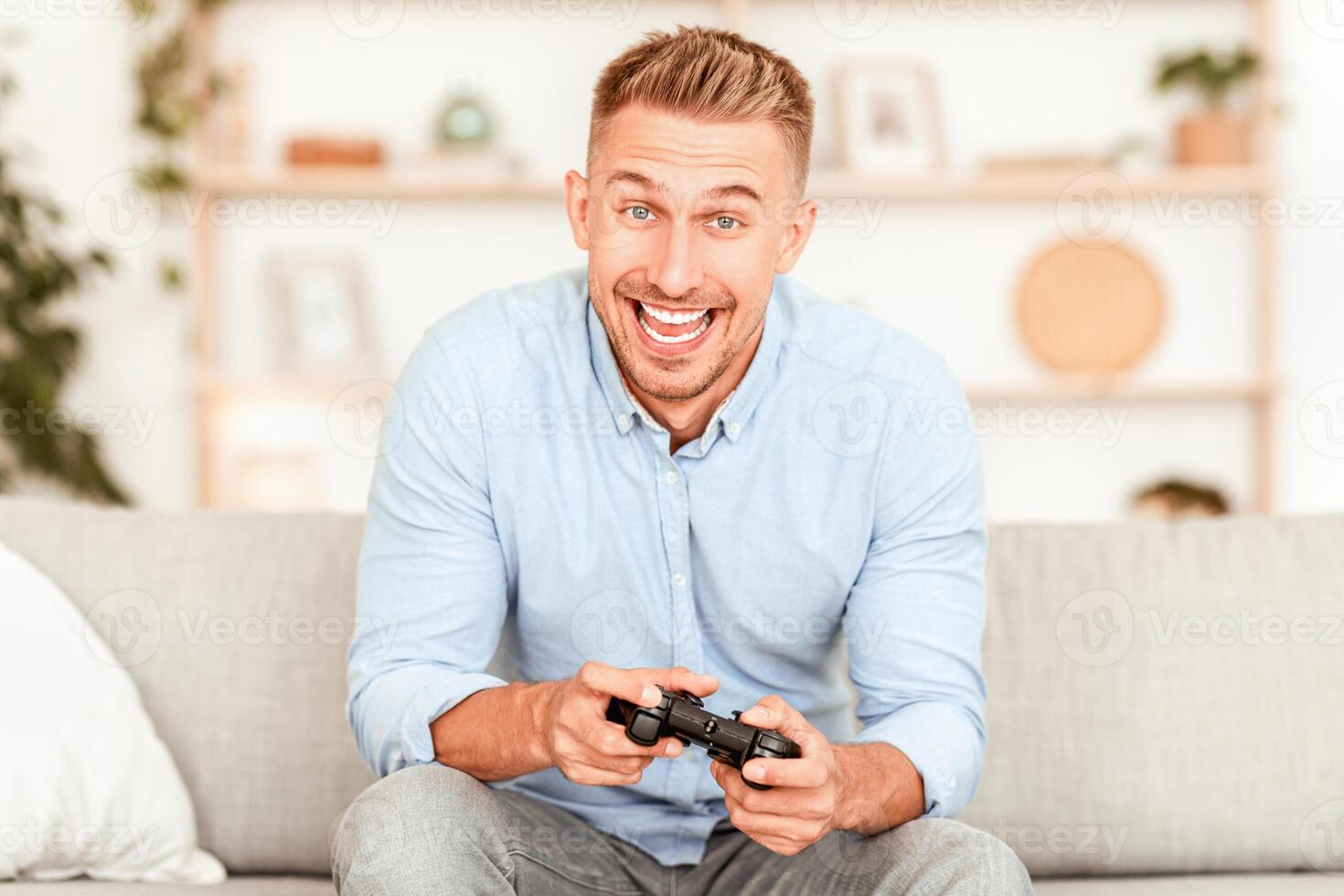 Online Competition Concept. Portrait of excited young man playing games, holding console joystick. Smiling adult gamer sitting on the couch at home in living room, copy space, blurred background photo