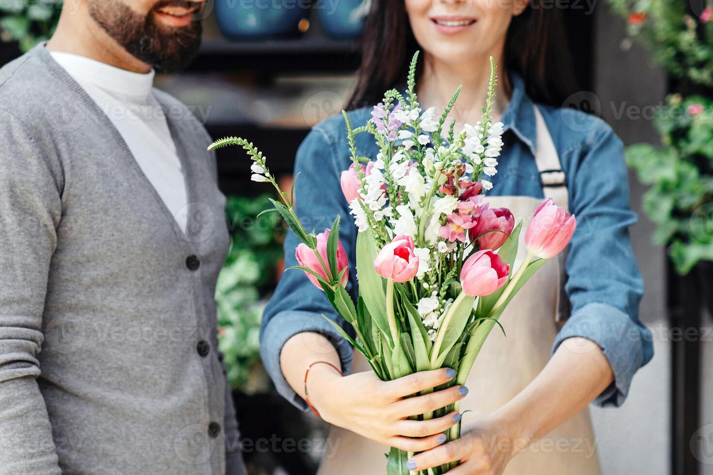 Blooming season, small business, flower studio and work of florist with client. Cheerful millennial european lady seller in apron showing fresh bouquet to man, plants store background, copy space photo