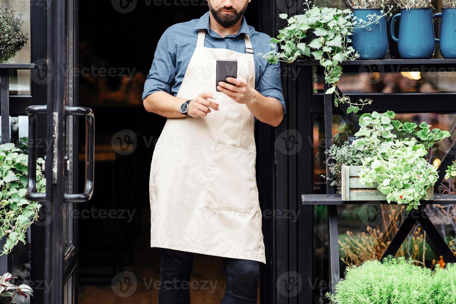 Online ordering of bouquet, selling plants in rustic floral studio. Serious young attractive bearded man in apron typing in smartphone in shop outdoor, with vases and different flowers in pots photo