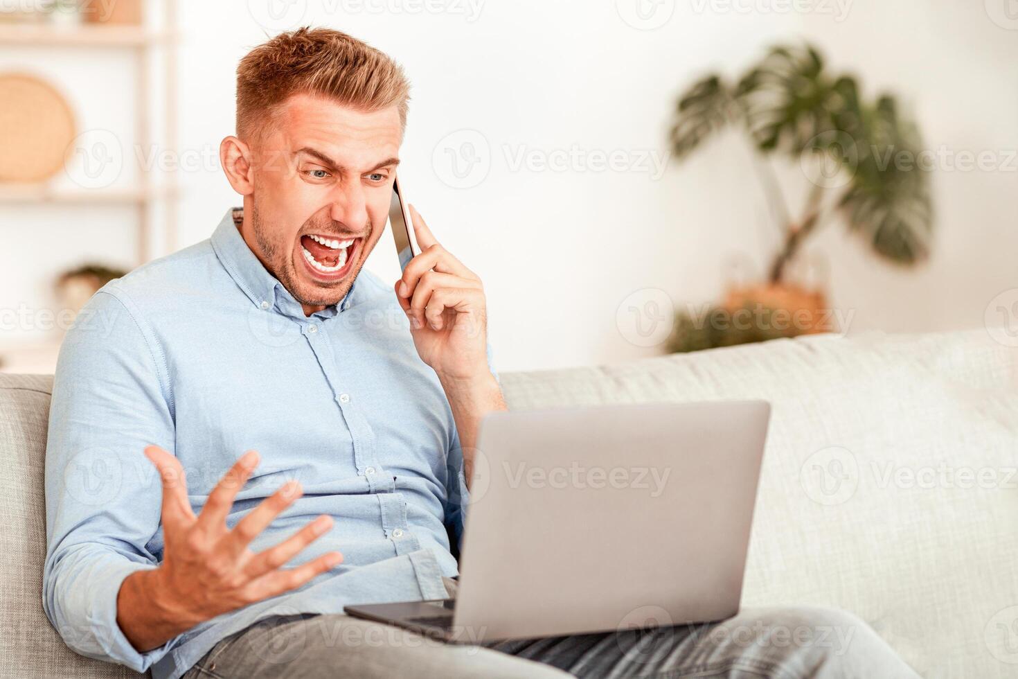 Angry irritated guy talking on mobile phone and shouting, sitting on sofa, using laptop, copy space photo