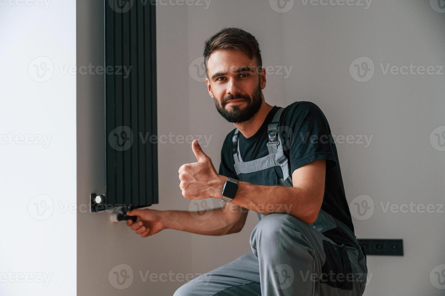 Sitting and showing ,thumb up. Plumber worker ,installing heating radiator in empty room of a newly built house photo