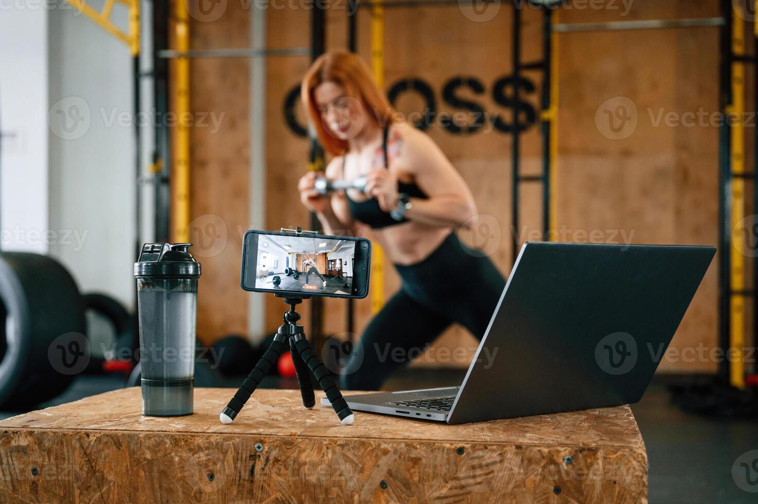 Laptop, bottle and smartphone on tripod is on the table. Beautiful strong woman is in the gym photo
