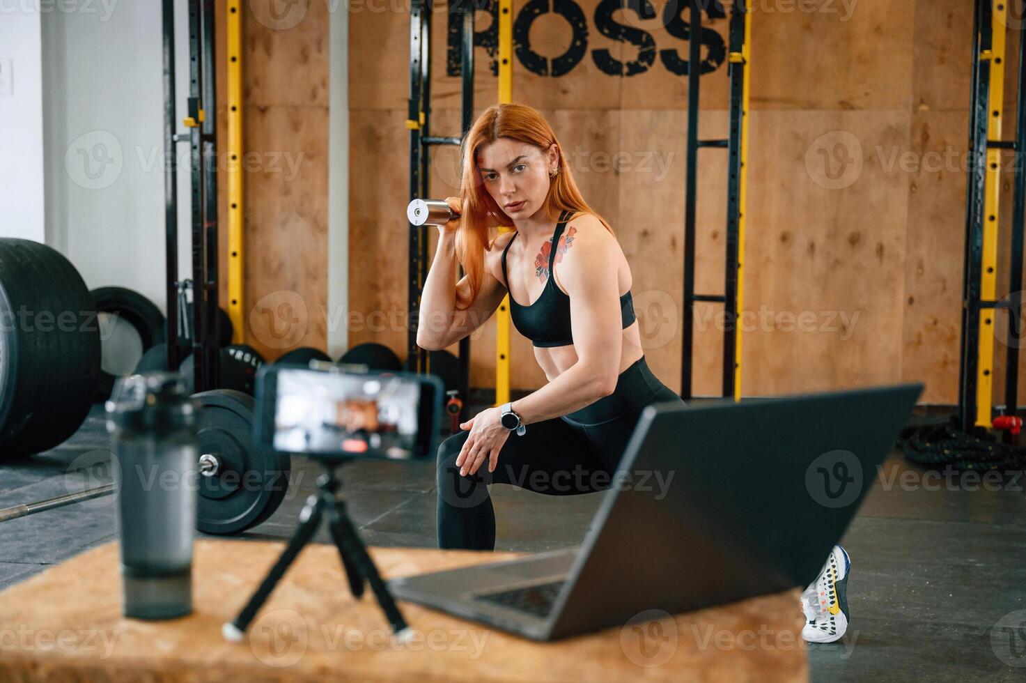 Laptop, bottle ,and smartphone on tripod is on the table. Beautiful strong woman is in the gym photo