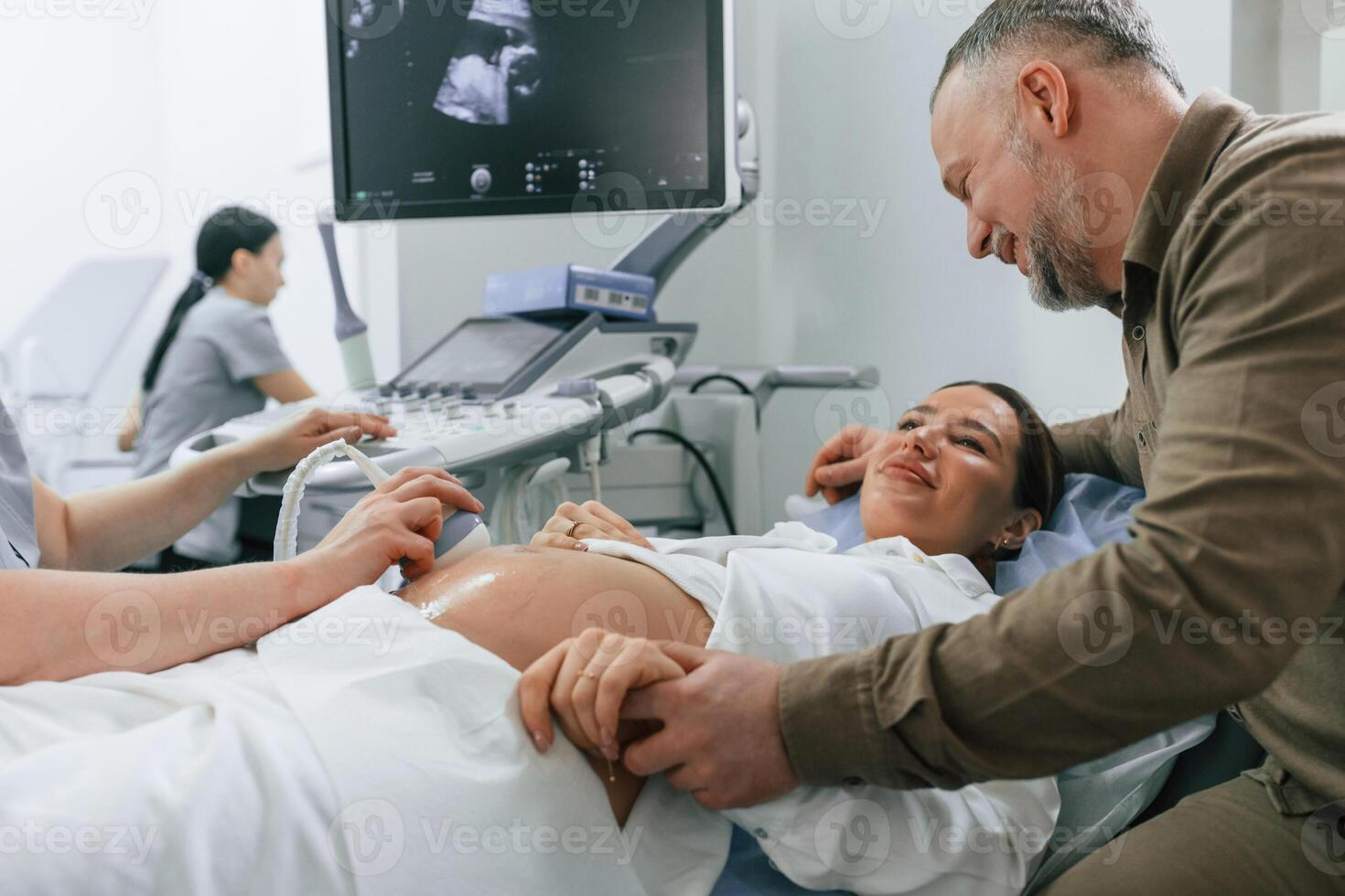 Happy husband is sitting ,and supporting his wife. Pregnant woman is lying down in the hospital, doctor does ultrasound photo