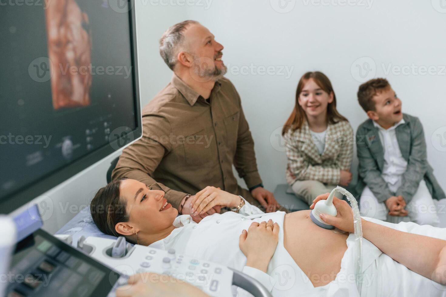 Husband, and, kids are sitting together. Pregnant woman is lying down in the hospital, doctor does ultrasound photo
