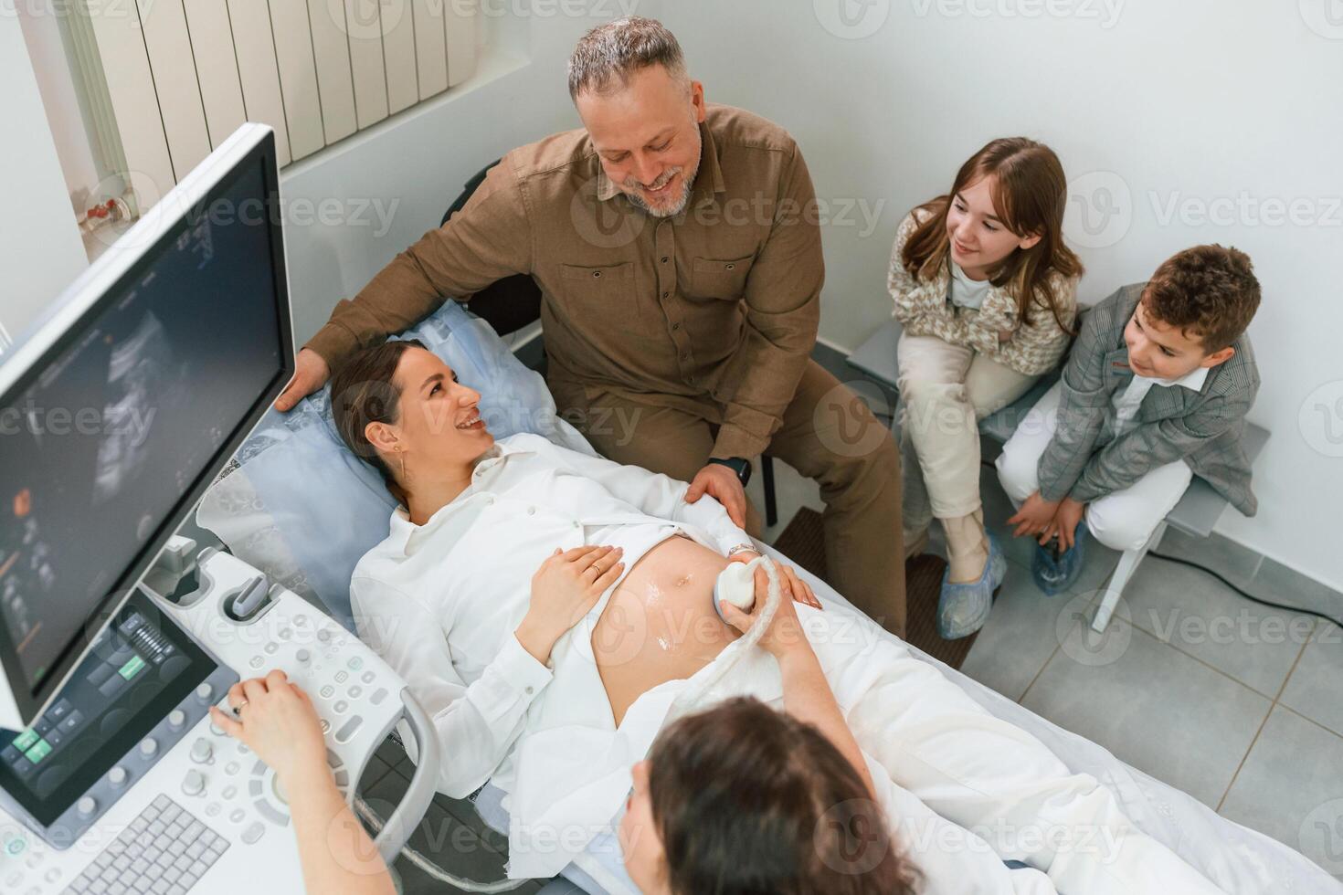 Top view. Kids and, husband supporting. Pregnant woman is lying down in the hospital, doctor does ultrasound photo