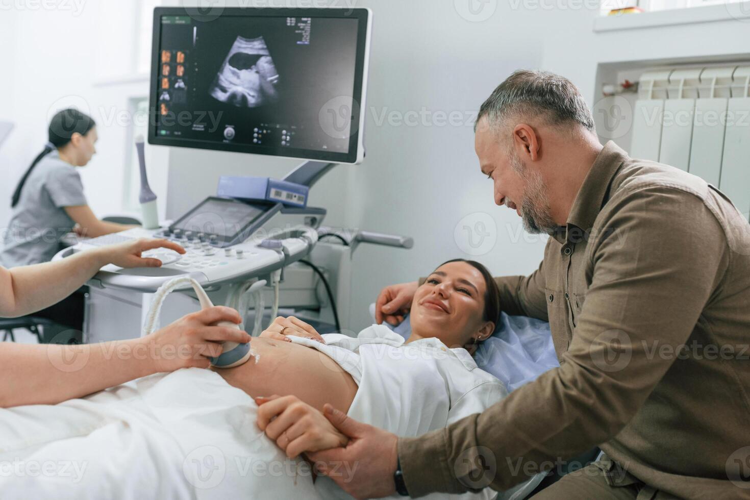 Husband is supporting, his wife. Pregnant woman is lying down in the hospital, doctor does ultrasound photo