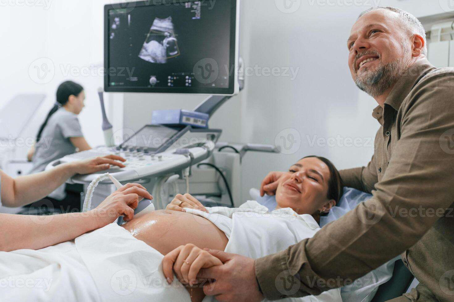 Happy, husband is sitting and supporting his wife. Pregnant woman is lying down in the hospital, doctor does ultrasound photo