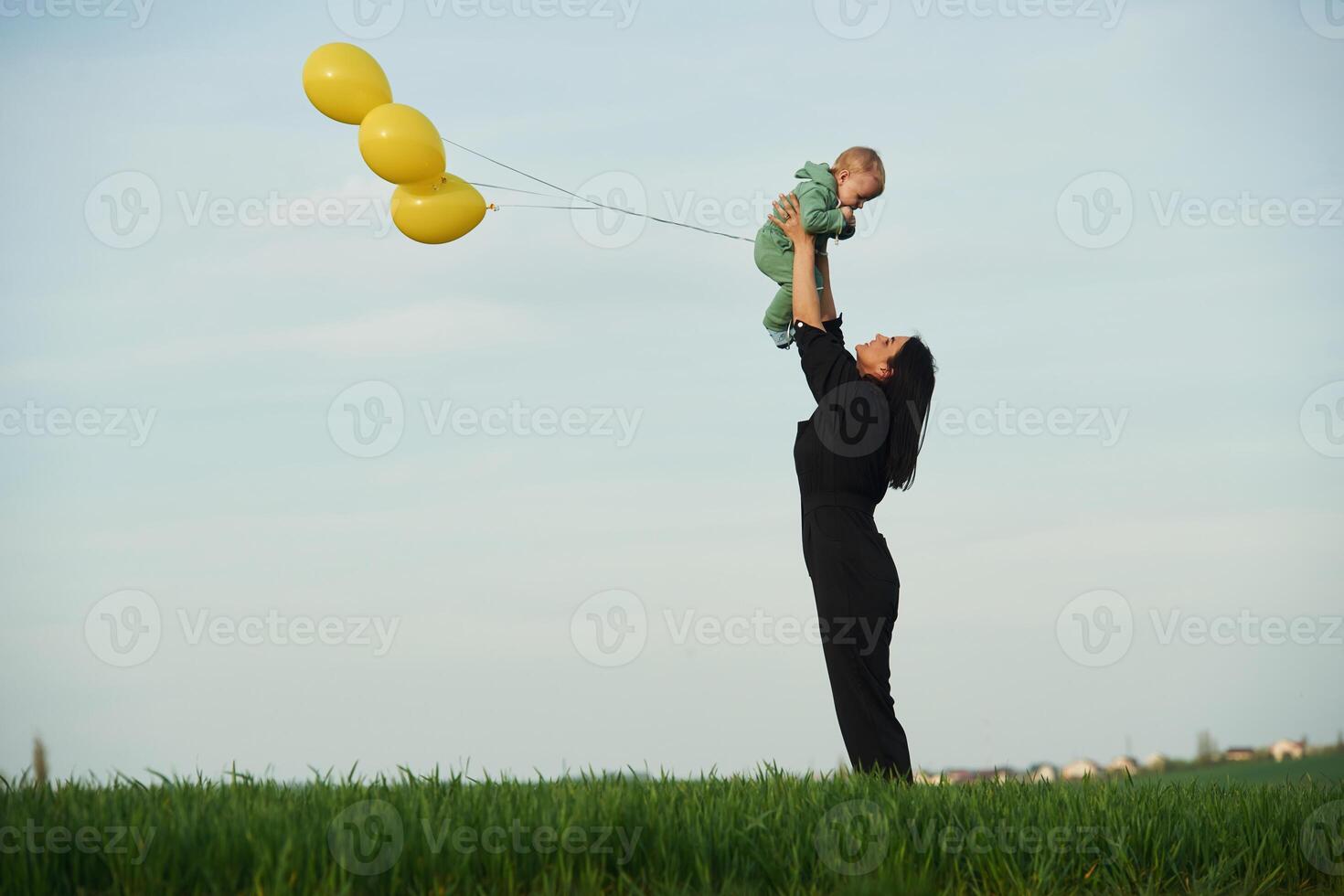 Yellow colored, balloons in hands. Mother with her little baby son is outdoors on the agricultural field photo