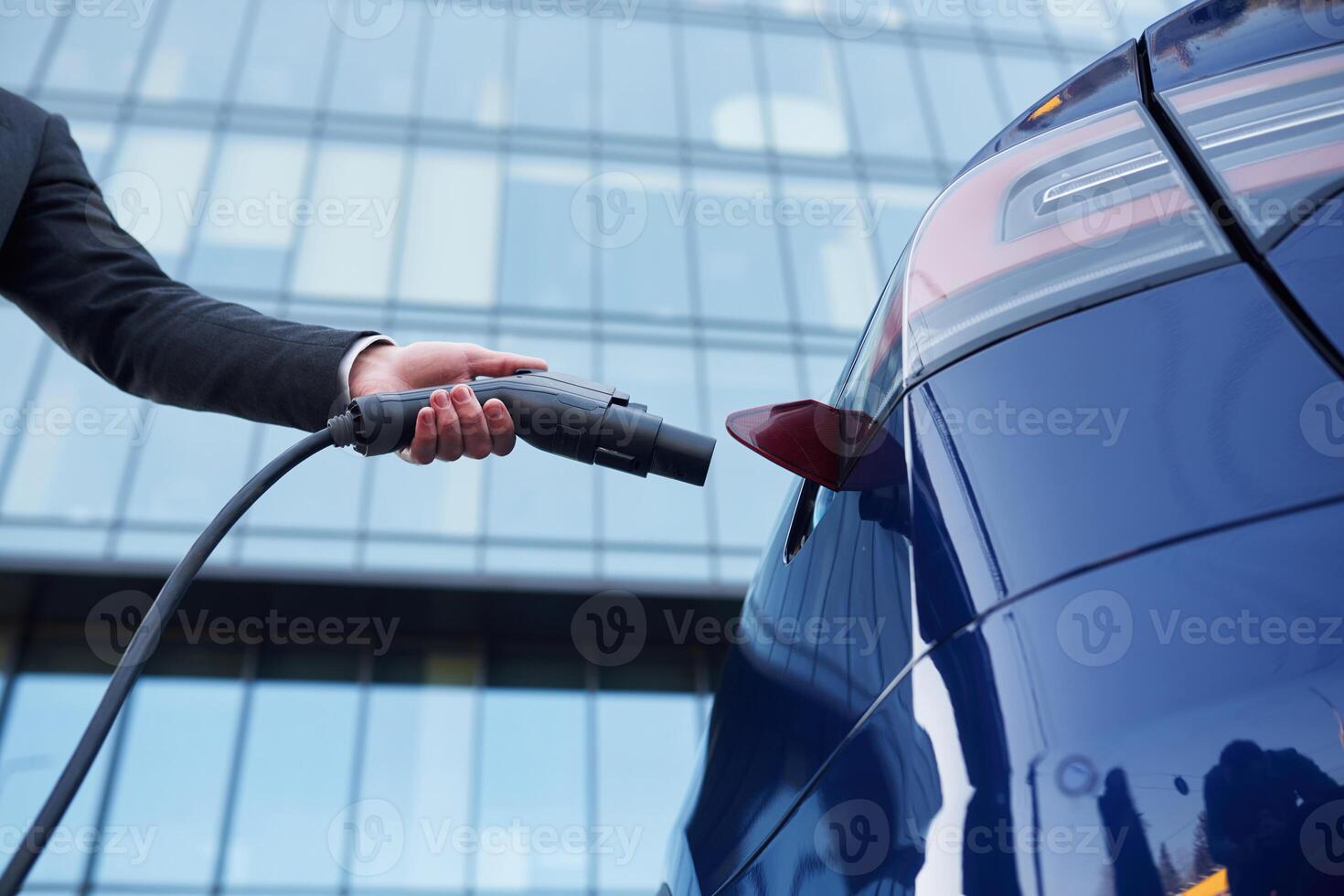 Close up view, putting, charger into the socket. Businessman is standing near his electric car outdoors photo