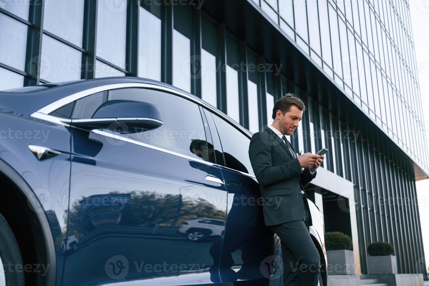 Holding smartphone.,. Businessman ,is standing near his car outdoors photo