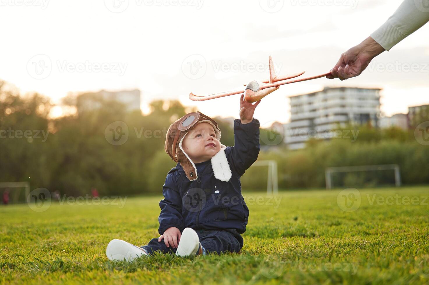 Little boy in aviator glasses is sitting on the field. Woman's hand giving toy plane to him photo