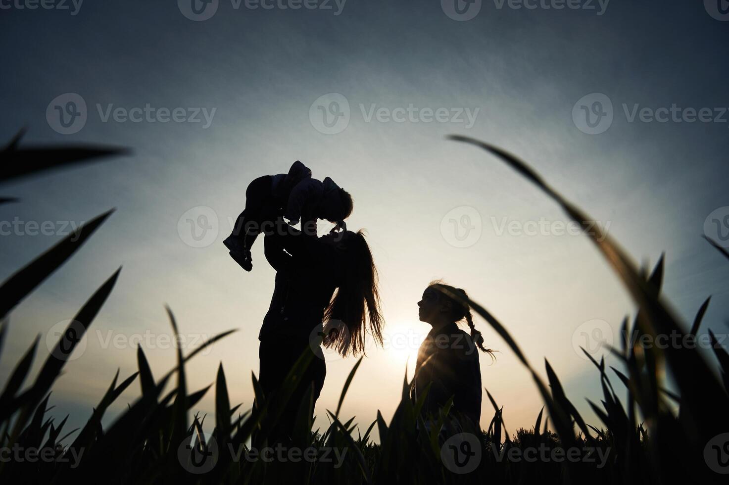 Silhouette,, of woman that holding her daughter in hands. Another girl is standing and watching photo