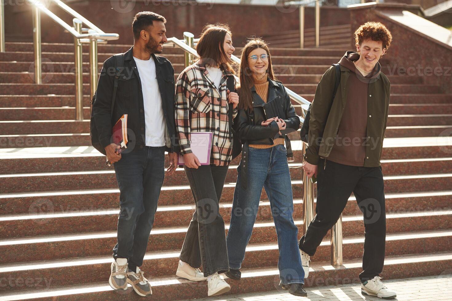 Walking, holding notepads. Four young ,,students in casual clothes are together outdoors photo