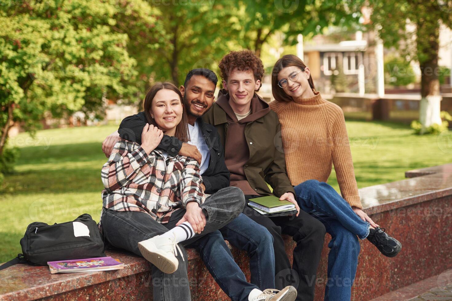Sitting and smiling,. Four young students in casual clothes are together outdoors photo