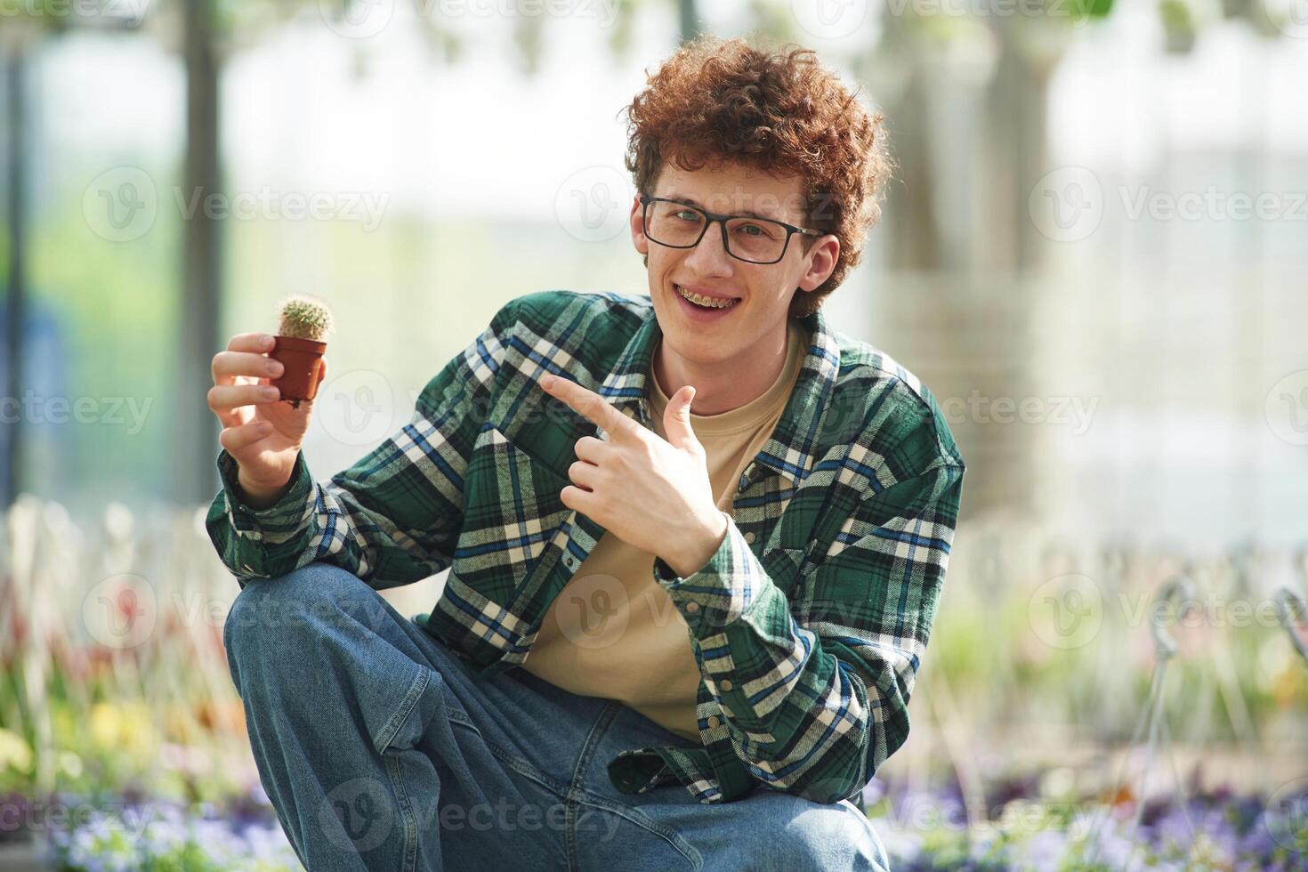 Sitting and holding the little cactus. Young man with curly hair and in glasses is in greenhouse photo