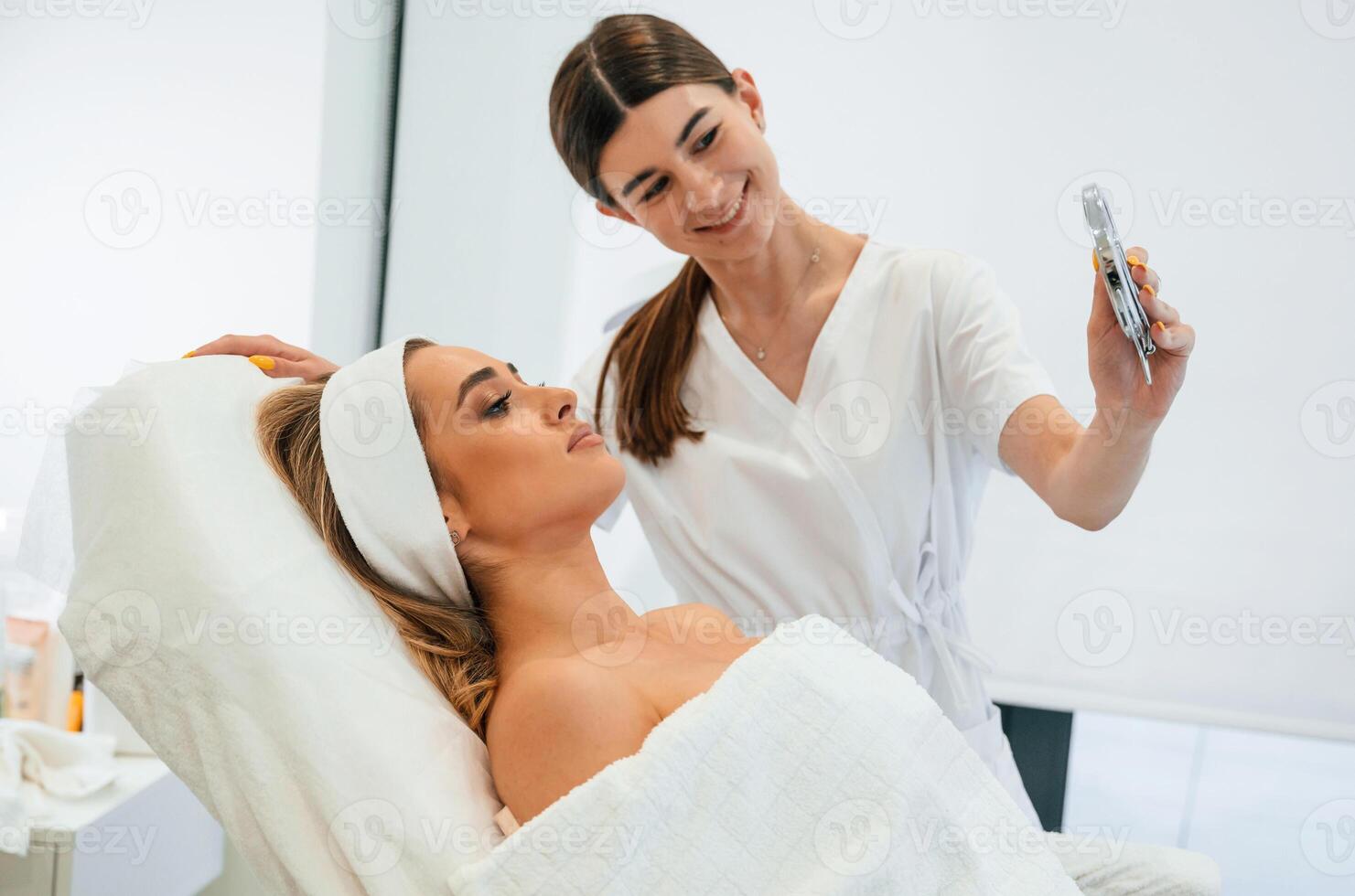 Holding, a mirror,, looking at herself. Woman face getting facial care by beautician at spa salon photo