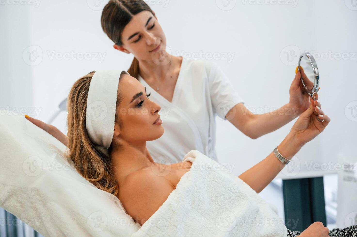 Holding, a mirror, looking ,at herself. Woman face getting facial care by beautician at spa salon photo