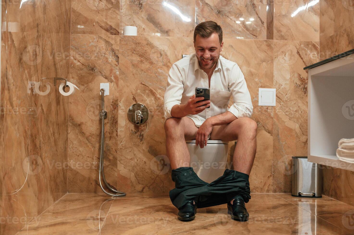 Watching funny content. A man in a white shirt is sitting on the toilet, holding a smartphone in his hands photo
