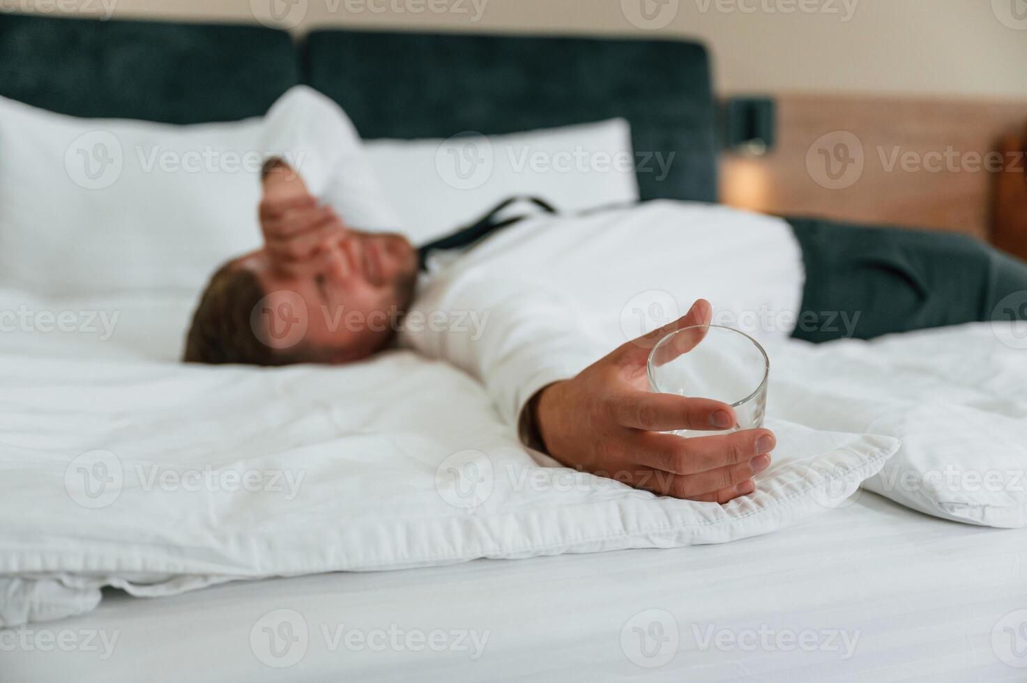 Empty glass in, hand, lying down on the bed, tired. Businessman is indoors in the hotel room photo