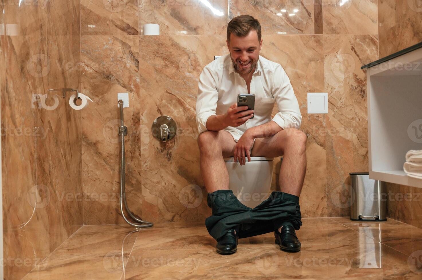 Watching funny content.. A man in a white shirt is sitting on the toilet, holding a smartphone in his hands photo