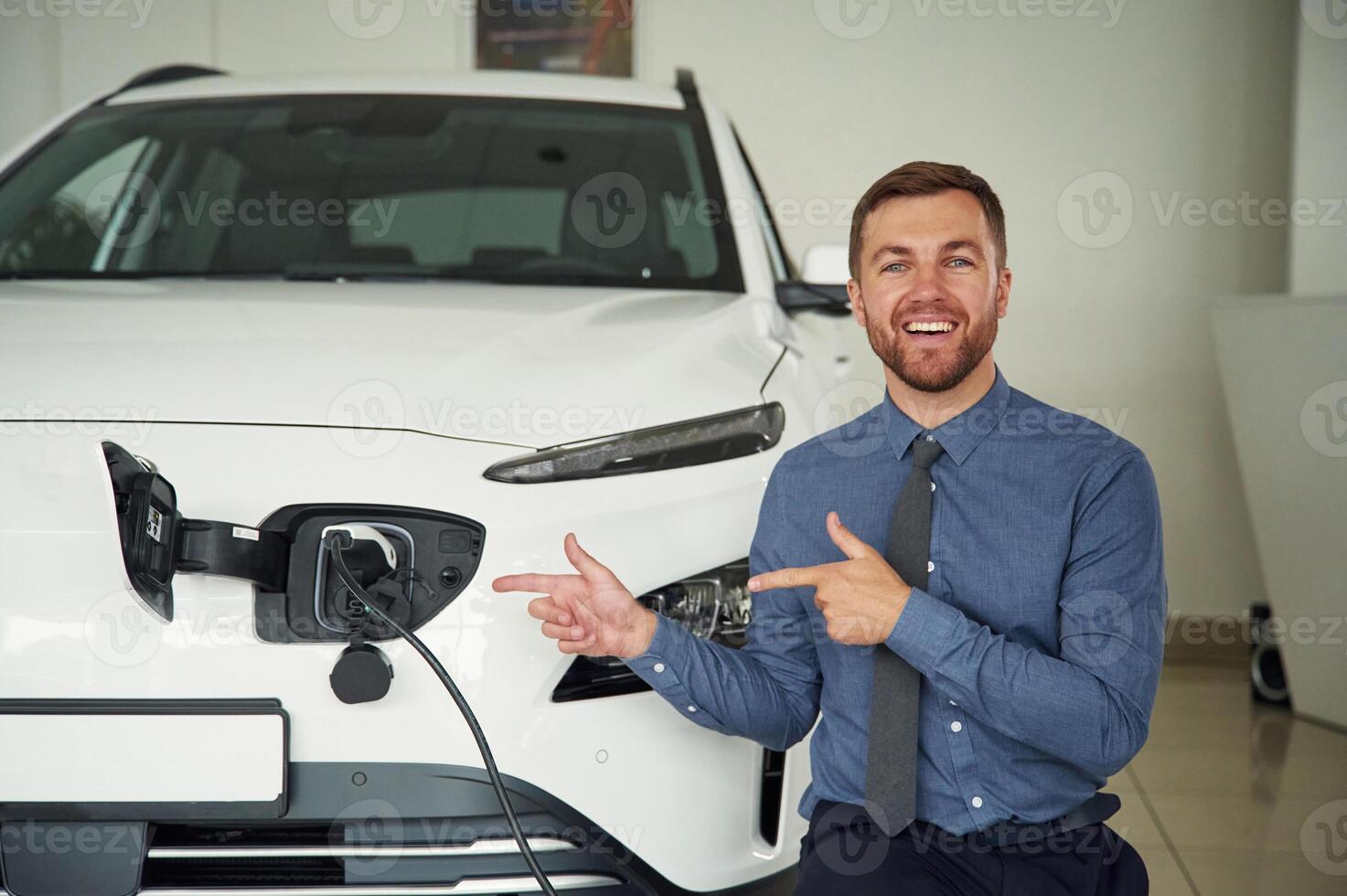 Showing, the charging process. Handsome car dealership worker is with electric vehicle photo